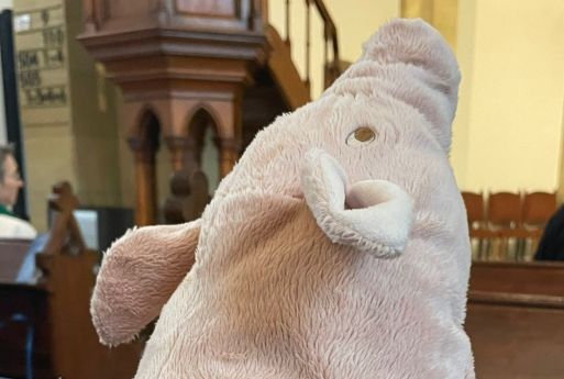 Foto: Im Vordergrund sehen wir den Kopf eines rosa-farbenen Plüsch-Schweins in einer Kirchenbank sitzend. im Hintergrund sehen wir links die Pfarrerin auf der ersten Kirchenbank. Am rechten Bildrand eine weitere Person und vorn eine Frau am Lesepult.