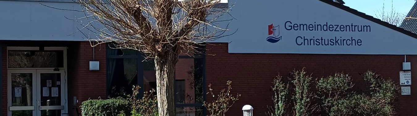 Foto: Ansicht vom Kirchhof auf das Kindergartengebäude des Gemeindezentrums an der Sandstraße. Linker Bildrand der Seiteneingang zum Gebäude, rechtes Bilddrittel das Logo und der Name des Gemeindezentrums Christuskirche. In der Mitte ein kleiner Baum auf