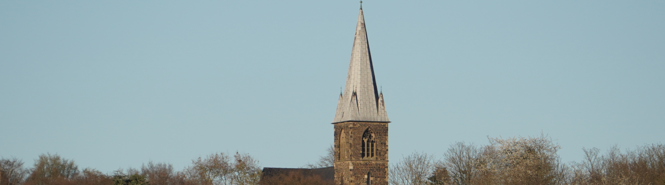 Foto des Kirchturms der Evangelischen Kirche der Trinitatis Kirchengemeinde in Witten. Blauer unbewölkter Himmel, am unteren Bildrand Baumkronen