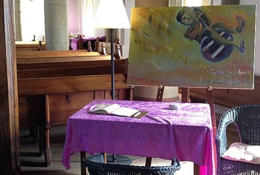 Foto: In der Kirche. Unmittelbar nach dem Eintritt in die Kirche fehlen auf der rechten Seite einige Kirchenbänke. Dort steht ein Tisch mit einem Gästebuch, ein Stuhl, ein großer Kerzenständer. Manchmal auch zusätzliche Deko. Hier eine Staffelei.
